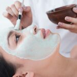 Eine Person trägt mit einem Pinsel eine grüne Gesichtsmaske auf das Gesicht einer Frau in einem Wellnessbereich auf.