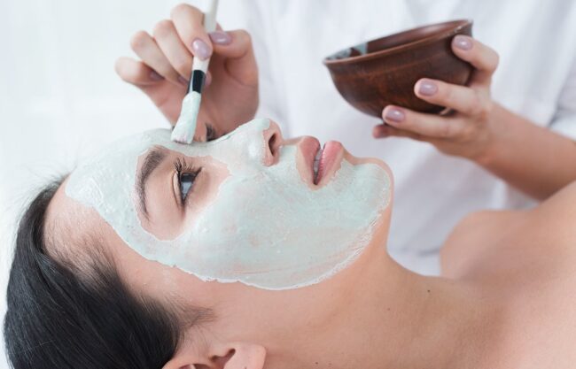 Eine Person trägt mit einem Pinsel eine grüne Gesichtsmaske auf das Gesicht einer Frau in einem Wellnessbereich auf.