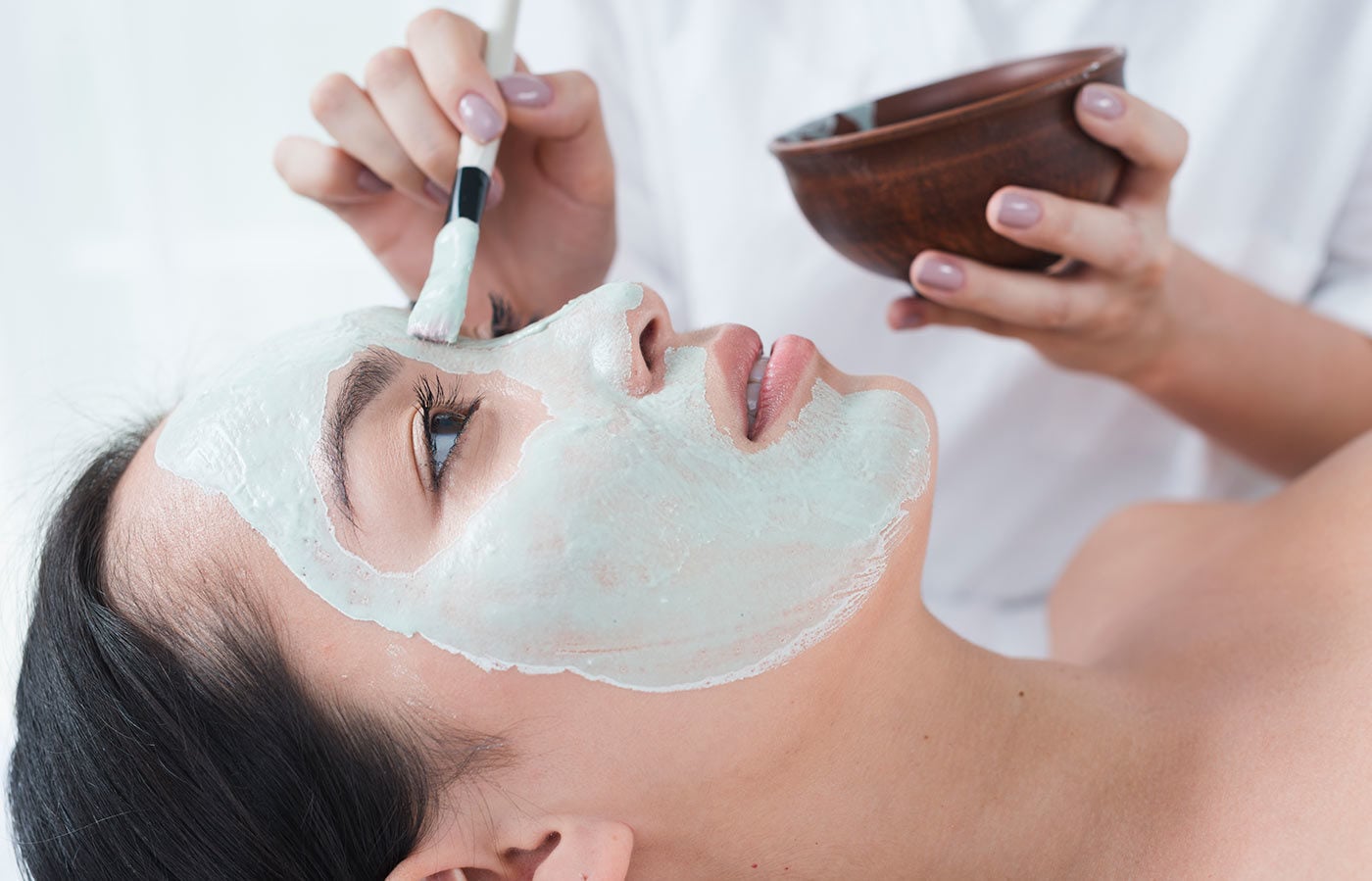 Eine Person trägt mit einem Pinsel eine grüne Gesichtsmaske auf das Gesicht einer Frau in einem Wellnessbereich auf.