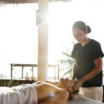 Eine Person genießt eine verjüngende Kopfmassage unter freiem Himmel von einem Therapeuten, umgeben von Sonnenlicht und Palmen, die die perfekte Spa- und Wellness-Atmosphäre schaffen.