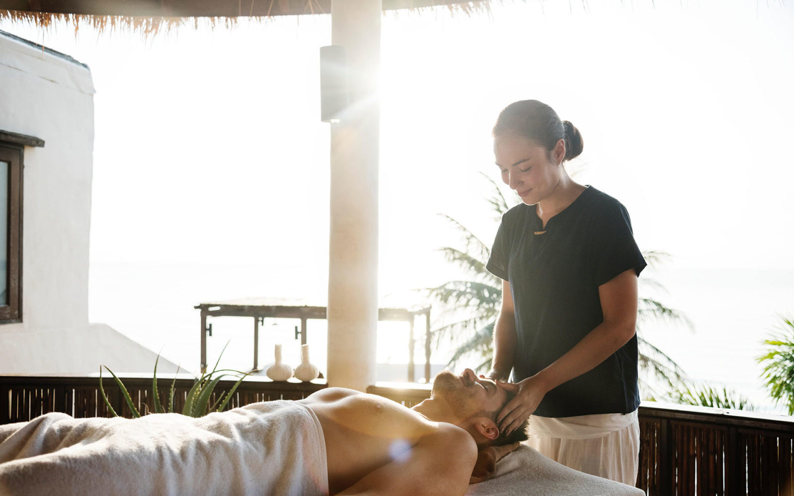 Eine Person genießt eine verjüngende Kopfmassage unter freiem Himmel von einem Therapeuten, umgeben von Sonnenlicht und Palmen, die die perfekte Spa- und Wellness-Atmosphäre schaffen.