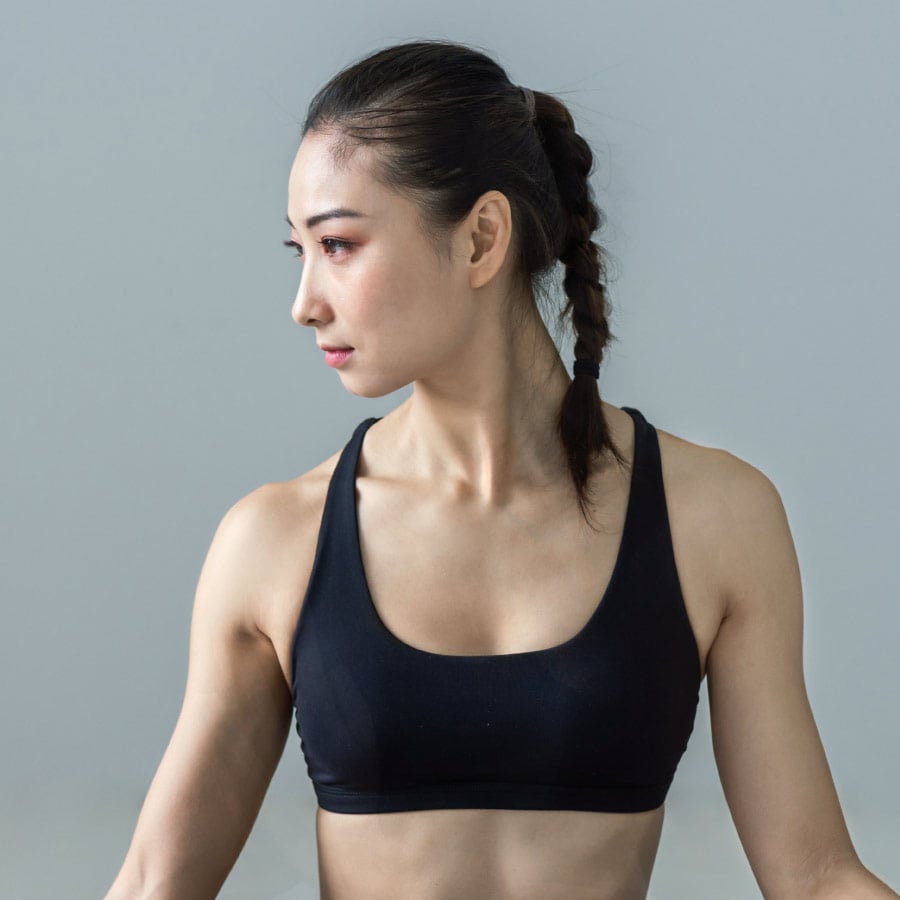Frau in einem schwarzen Leonardo-Sport-BH mit geflochtenem Haar blickt zur Seite vor einem schlichten Hintergrund.