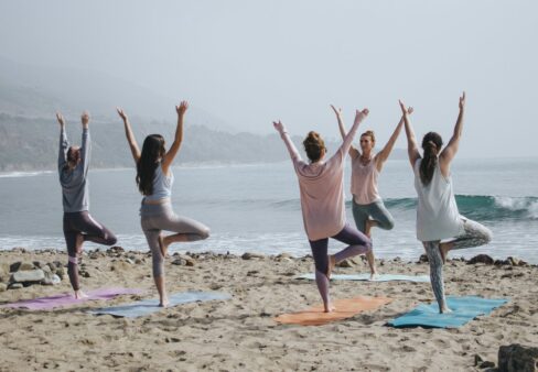 Fünf Personen, die auf Yogamatten an einem Sandstrand am Meer Yoga in der Baumstellung machen, mit Wellen im Hintergrund.