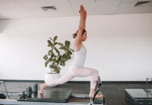 Frau in Sportkleidung macht einen Ausfallschritt mit erhobenen Armen auf einem Pilates-Reformer in einem hellen Studio.