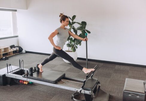 Frau bei einer Dehnungsübung auf einem Pilates-Reformer-Gerät in einem hellen Studio mit einer Pflanze im Hintergrund.
