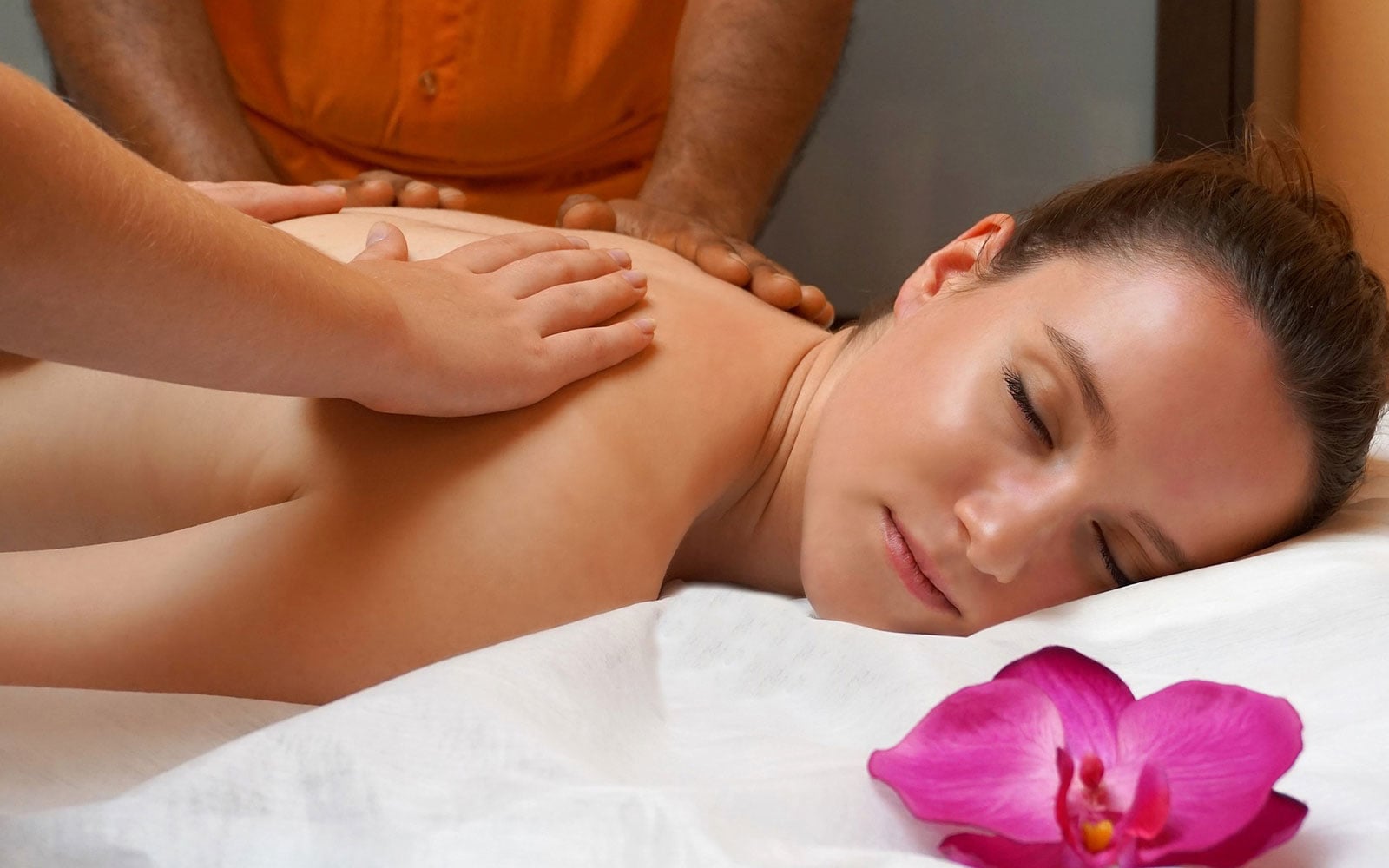 Frau liegt mit geschlossenen Augen auf einer Massageliege und erhält eine Rückenmassage, im Vordergrund eine rosa Orchidee.