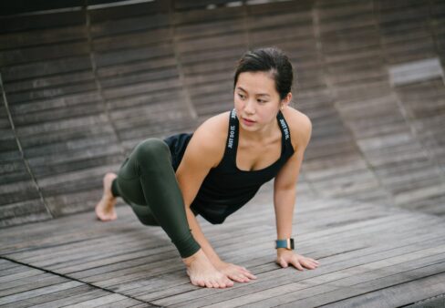 Frau, die sich auf einem Holzboden beim Yoga dehnt, trägt ein schwarzes Tanktop und grüne Leggings.