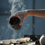 Eine Hand gießt dampfenden Tee aus einer Teekanne in kleine Tassen auf einem Tisch bei gedämpftem Licht.