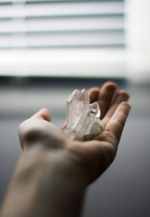 Eine Hand, die klare Quarzkristalle vor einem unscharfen Hintergrund mit Jalousien hält.