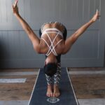 Frau in Yogastellung, nach vorne gebeugt mit weit ausgestreckten Armen auf einer Yogamatte in einem Raum.