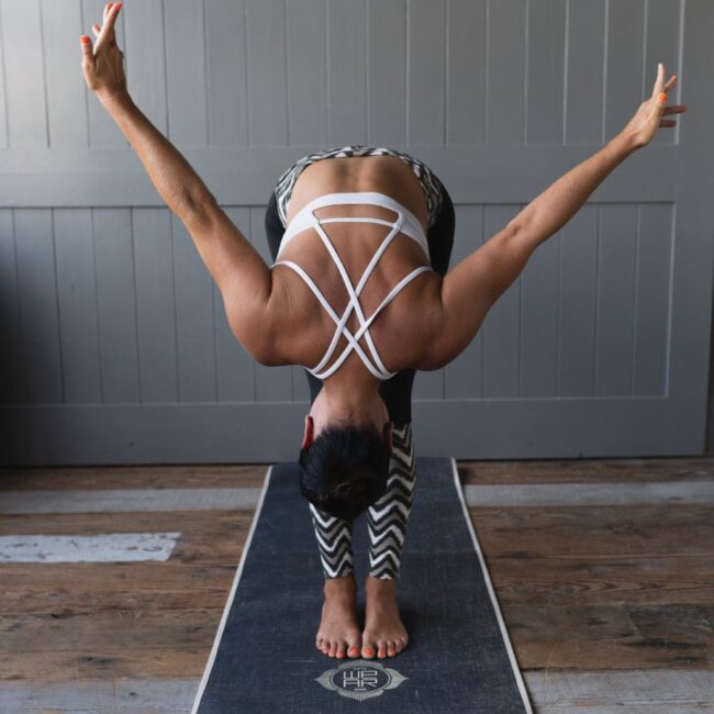 Frau in Yogastellung, nach vorne gebeugt mit weit ausgestreckten Armen auf einer Yogamatte in einem Raum.