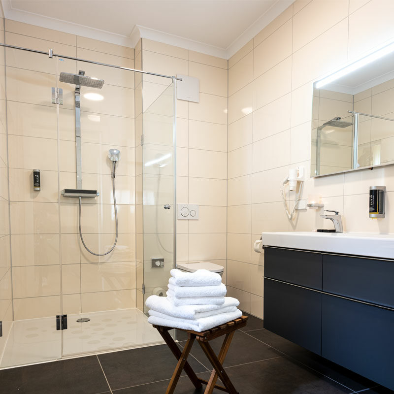 Modernes Badezimmer im Deluxe-Doppelzimmer mit Glasdusche, Doppelwaschbecken, Spiegel und einem Hocker, auf dem ordentlich gefaltete weiße Handtücher liegen.