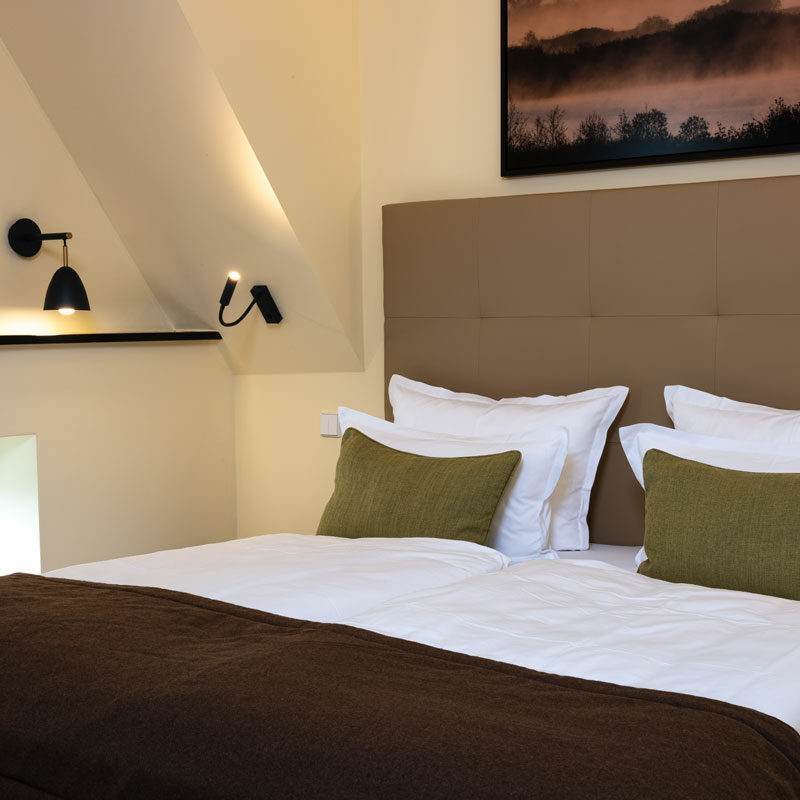 Modernes Deluxe Doppelzimmer mit ordentlich gemachtem Bett, grünen Kissen, Wandlampen und einem Landschaftsfoto über dem Kopfteil.