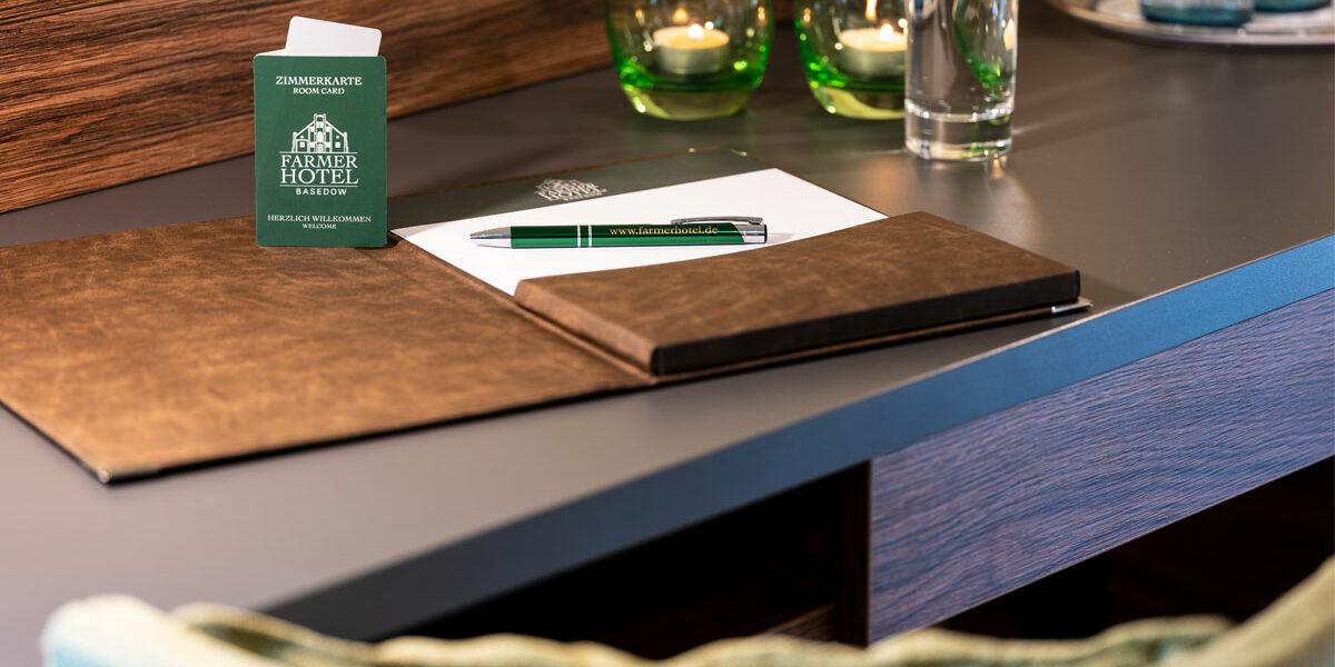 Ein Hoteltisch in einem Deluxe-Doppelzimmer mit einem Ordner, einem Stift, einem Notizblock, Kerzen, einem Glas Wasser und einer Zimmerschlüsselkarte.