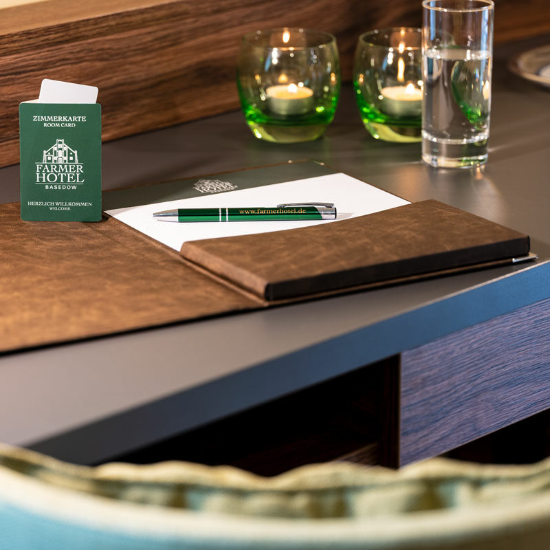 Ein Hoteltisch in einem Deluxe-Doppelzimmer mit einem Ordner, einem Stift, einem Notizblock, Kerzen, einem Glas Wasser und einer Zimmerschlüsselkarte.