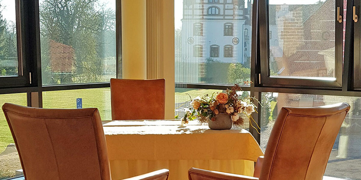 Zwei Ledersessel an einem blumengeschmückten Tisch mit Blick durch große Fenster auf einen Garten und ein weißes historisches Gebäude.