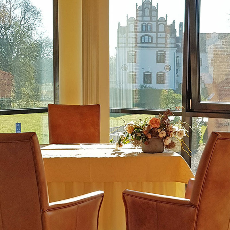 Zwei Ledersessel an einem blumengeschmückten Tisch mit Blick durch große Fenster auf einen Garten und ein weißes historisches Gebäude.