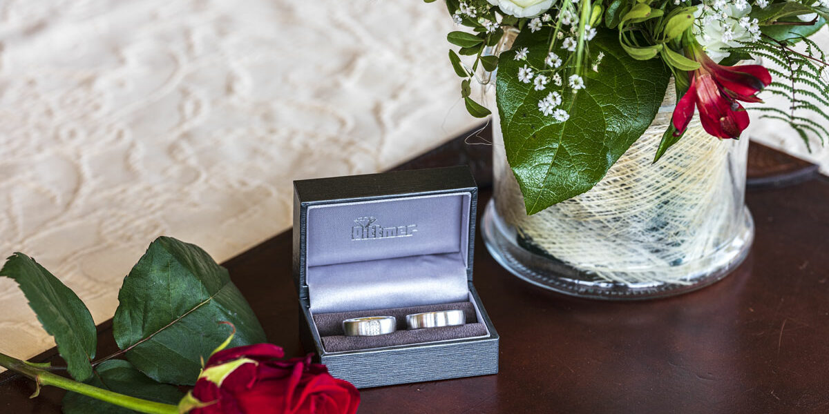 Zwei Eheringe in einer Schachtel neben einer roten Rose und einem Blumenstrauß auf einem Tisch.