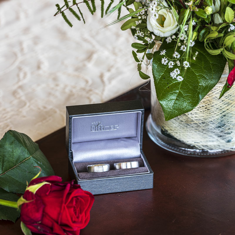 Zwei Eheringe in einer Schachtel neben einer roten Rose und einem Blumenstrauß auf einem Tisch.