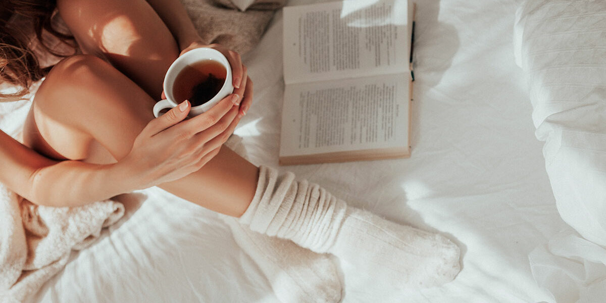 Eine Person in gemütlichen Socken sitzt mit einer Tasse Tee in der Hand neben einem aufgeschlagenen Buch im sanften Sonnenlicht auf einem Doppelbett.