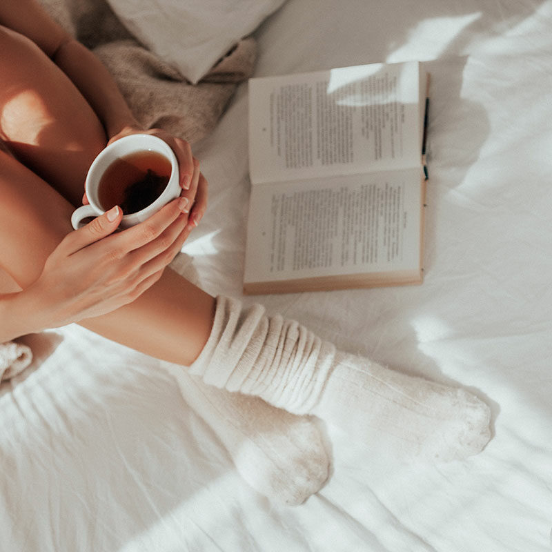 Eine Person in gemütlichen Socken sitzt mit einer Tasse Tee in der Hand neben einem aufgeschlagenen Buch im sanften Sonnenlicht auf einem Doppelbett.