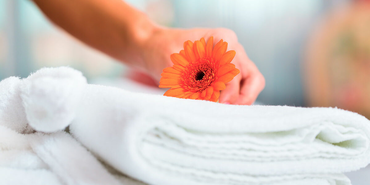 Eine Hand legt ein orangefarbenes Gänseblümchen auf ordentlich gefaltete weiße Handtücher in einem gemütlichen Doppelzimmer, was auf ein Spa oder eine entspannende Umgebung hindeutet.