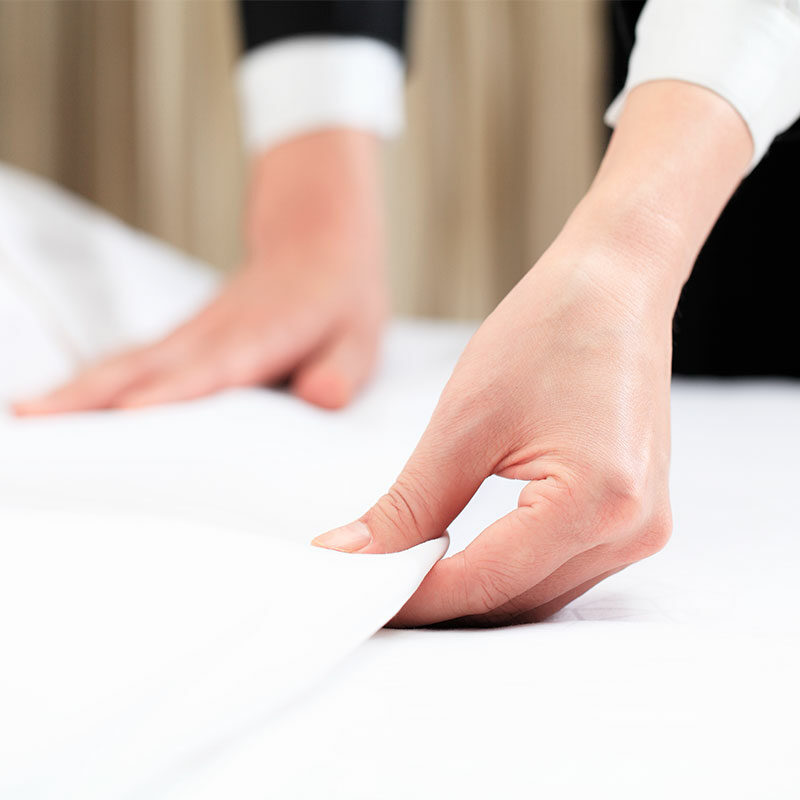 Nahaufnahme der Hände einer Person, die ein weißes Bettlaken auf einem Doppelbett ordentlich zurechtrückt.