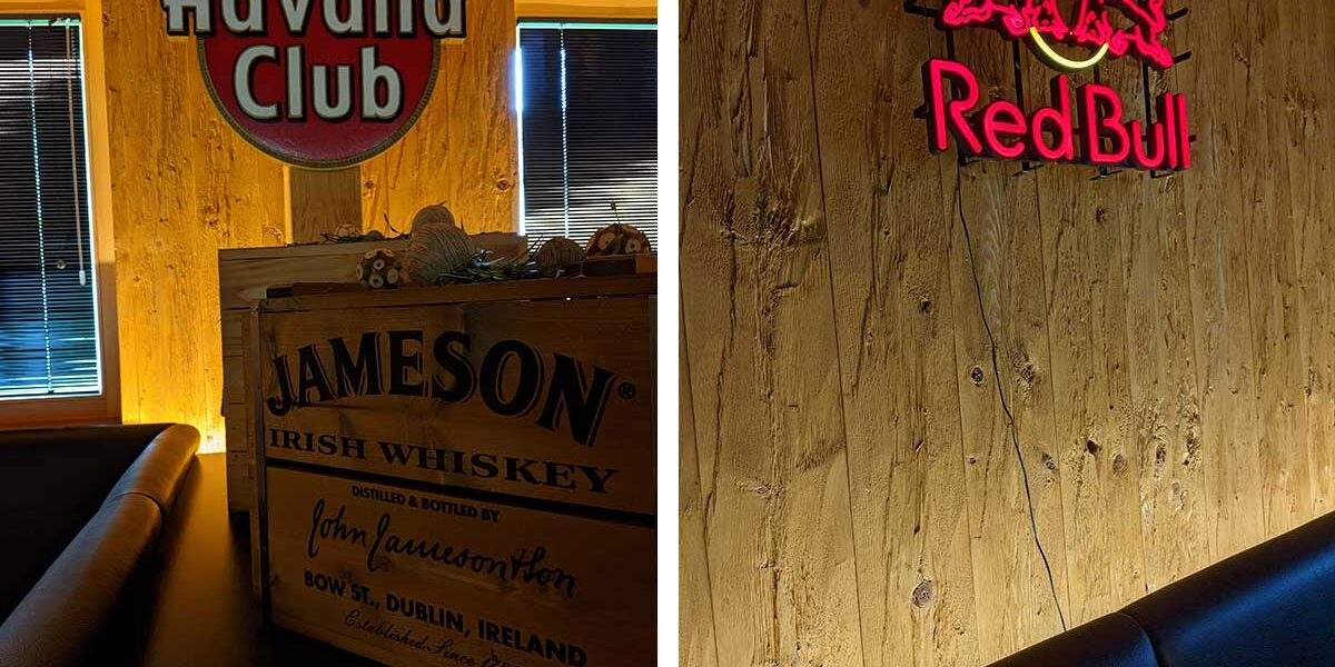 Links: Havana Club und Jameson-Schilder; Rechts: Red Bull-Leuchtreklame an einer Holzwand in einer schummrigen Bar im Stil eines Doppelzimmer.