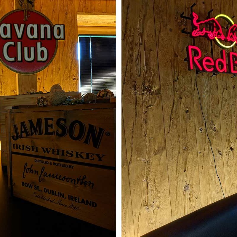 Links: Havana Club und Jameson-Schilder; Rechts: Red Bull-Leuchtreklame an einer Holzwand in einer schummrigen Bar im Stil eines Doppelzimmer.