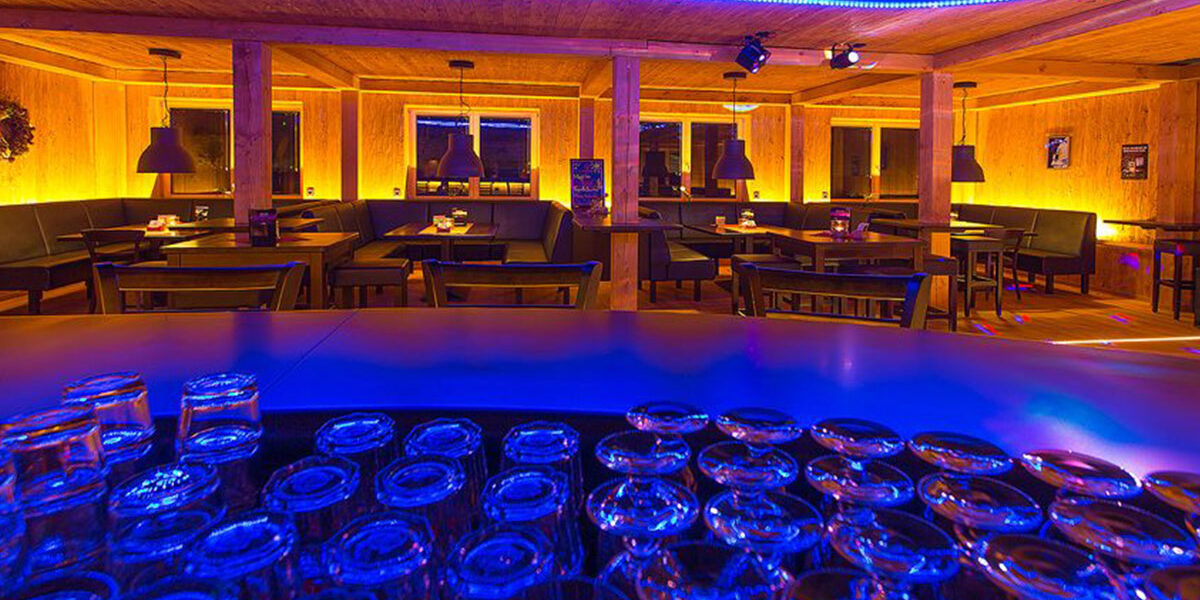 Das Innere der Bar im Kleiner Lord mit leeren Tischen, leuchtenden gelben Lichtern und auf dem Tresen gestapelten Gläsern bei blauer Beleuchtung.