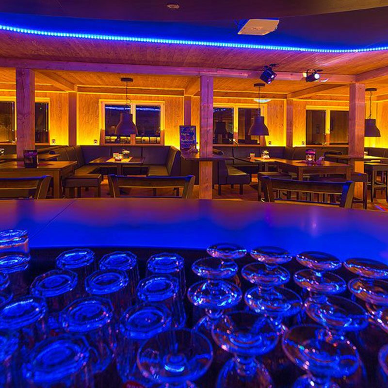 Das Innere der Bar im Kleiner Lord mit leeren Tischen, leuchtenden gelben Lichtern und auf dem Tresen gestapelten Gläsern bei blauer Beleuchtung.