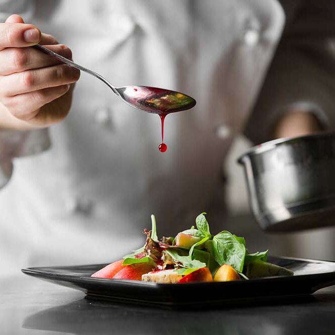 Ein Koch träufelt mit einem Löffel Sauce über einen angerichteten Salat und bereitet so ein optisch ansprechendes Gericht zu.