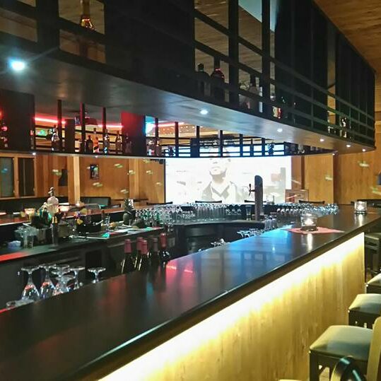 Moderne Bar mit Holzdekor, hohen Stühlen, Flaschenregalen und einem großen Bildschirm im Hintergrund - perfekt, um einen Kleinen Lord zu genießen.