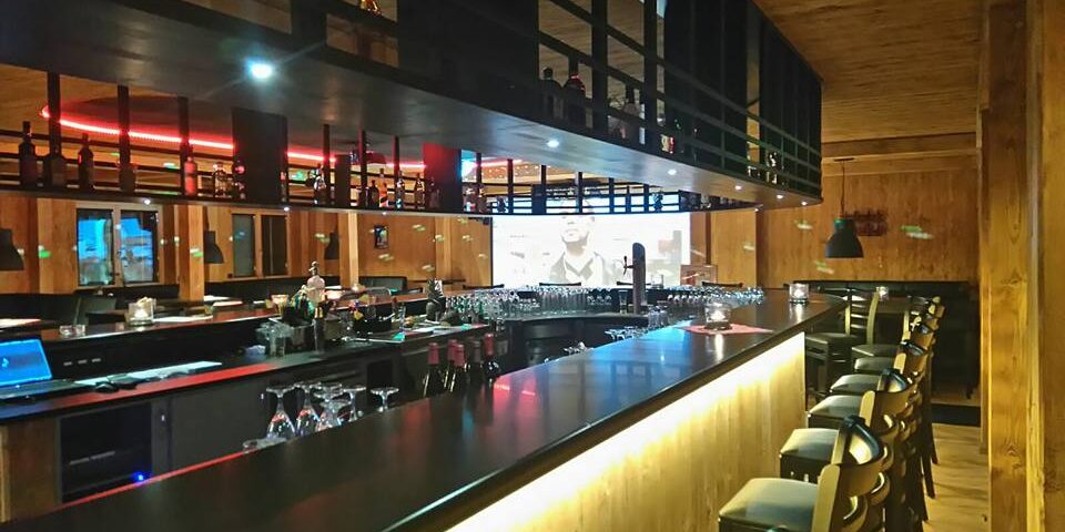 Moderne Bar mit Holzdekor, hohen Stühlen, Flaschenregalen und einem großen Bildschirm im Hintergrund - perfekt, um einen Kleinen Lord zu genießen.