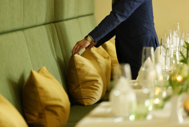 Eine Person in einem blauen Anzug rückt ein gelbes Kissen auf einer grünen Bank in einem Restaurant zurecht und bereitet sich auf ein angenehmes Essen- oder Trinkerlebnis vor.