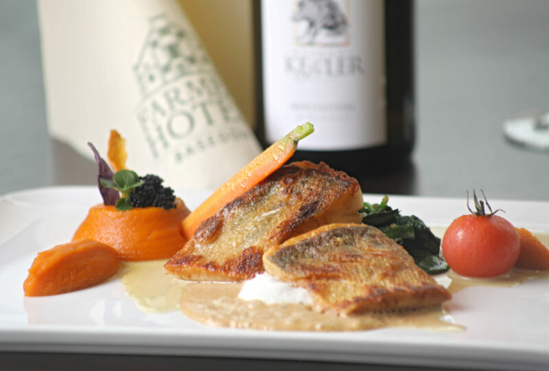 Gebratene Fischfilets mit Karottenpüree, Spinat, einer Kirschtomate und einer Flasche Weißwein im Hintergrund - perfekt für elegante Momente beim Essen und Trinken.