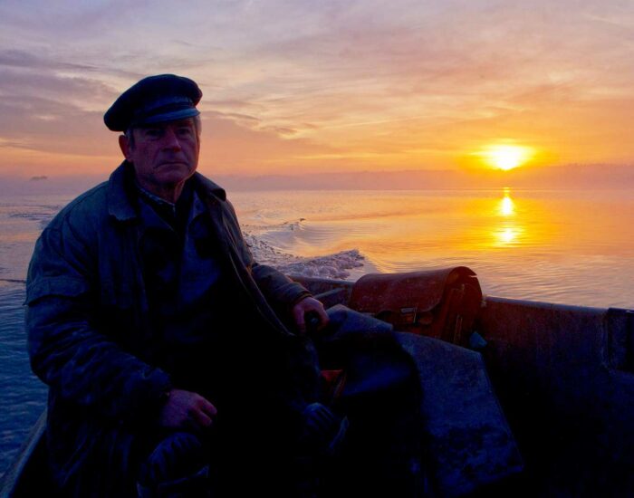 Älterer Mann mit Mütze sitzt in einem Boot auf ruhigem Wasser bei Sonnenuntergang, mit orangefarbenem Himmel im Hintergrund. Älterer Mann mit Mütze sitzt in einem Boot auf ruhigem Wasser bei Sonnenuntergang, mit orangefarbenem Himmel im Hintergrund.