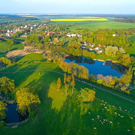 Luftaufnahme einer ländlichen Landschaft mit grünen Feldern, kleinen Seen, Bäumen und einem Dorf in der Ferne.