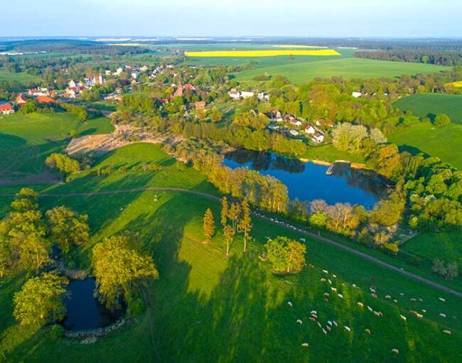 Luftaufnahme einer ländlichen Landschaft mit grünen Feldern, kleinen Seen, Bäumen und einem Dorf in der Ferne.