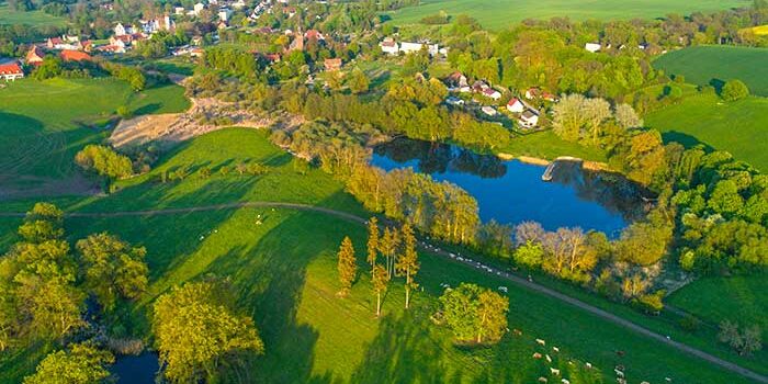 Luftaufnahme einer ländlichen Landschaft mit grünen Feldern, kleinen Seen, Bäumen und einem Dorf in der Ferne.