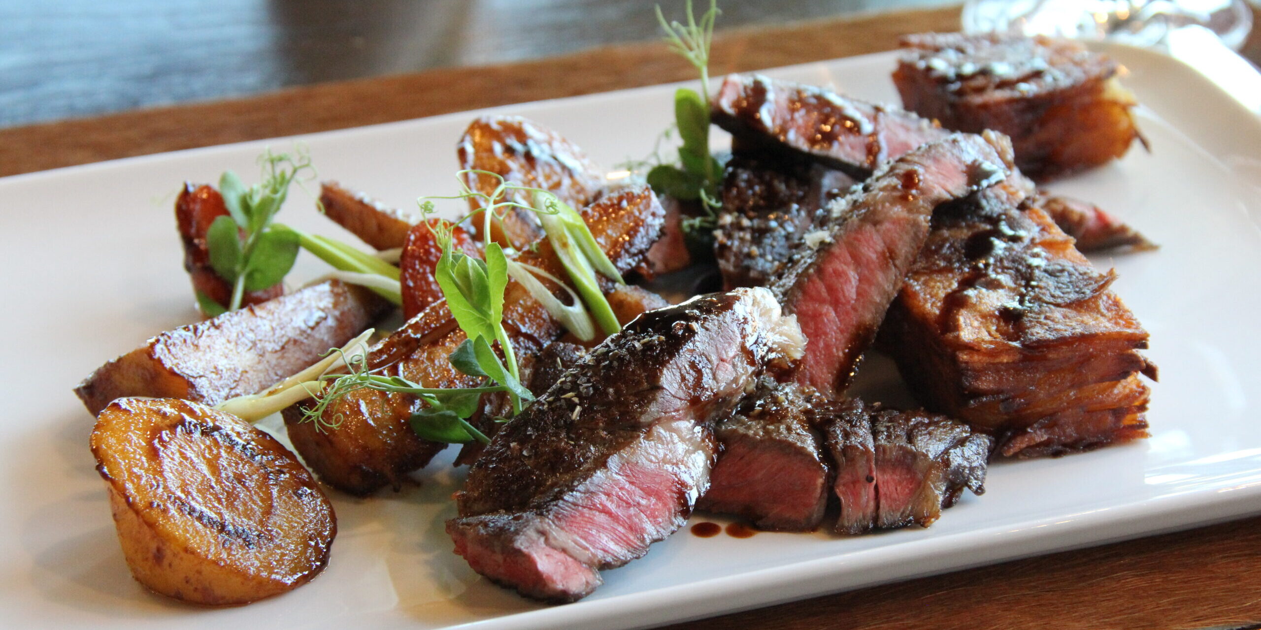 Geschnittenes Steak mit Bratkartoffeln und frischen Kräutern, fachmännisch zubereitet von unserem Koch (m/w/d), serviert auf einem weißen rechteckigen Teller.
