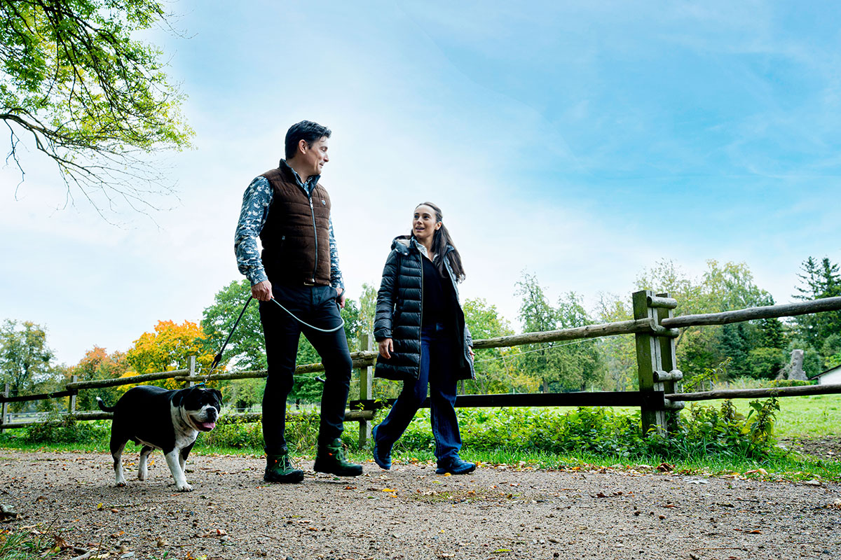 Ein Mann und eine Frau gehen gemeinsam mit ihrem Hund auf einem von Bäumen gesäumten Weg neben einem Holzzaun unter blauem Himmel spazieren.
