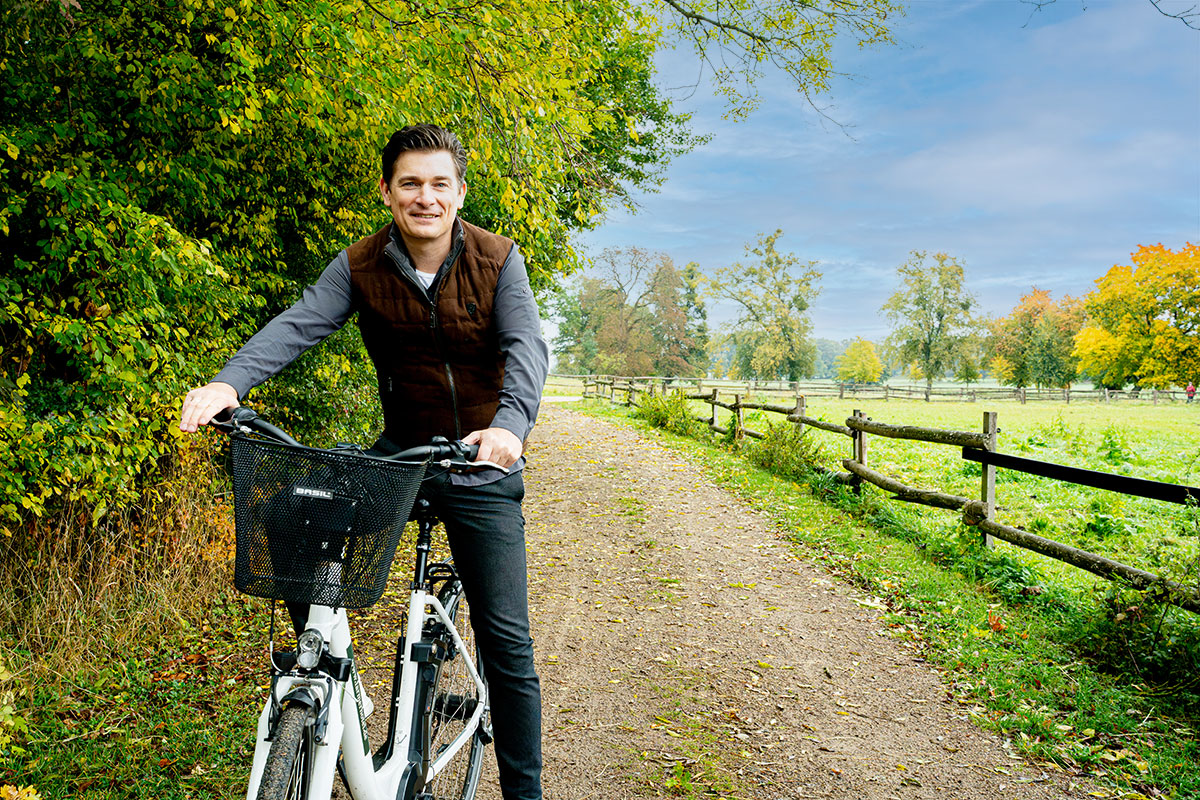 Ein Mann lächelt, während er mit seinem Fahrrad auf einem von Bäumen gesäumten Weg neben einem Holzzaun auf dem Land steht.