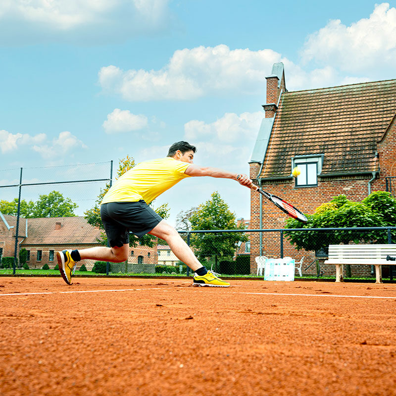 Mann in gelbem Hemd spielt Tennis auf einem Sandplatz und versucht, den Ball in der Nähe eines Backsteinhauses zu treffen.