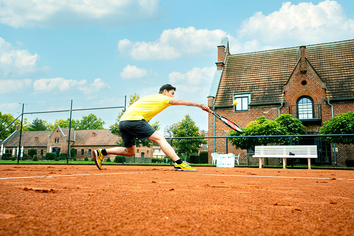 Mann in gelbem Hemd spielt Tennis auf einem Sandplatz und versucht, den Ball in der Nähe eines Backsteinhauses zu treffen.