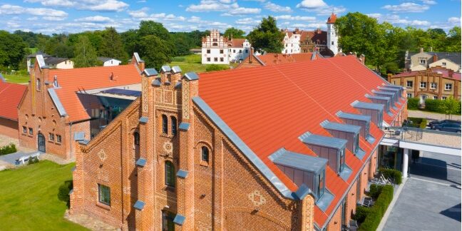 Ein Backsteingebäude mit rotem Dach und Dachgauben, umgeben von grünem Gras unter einem blauen Himmel mit Wolken – eine perfekte Kulisse für die Reittage, an denen jedes Pferd bereit ist, loszureiten.