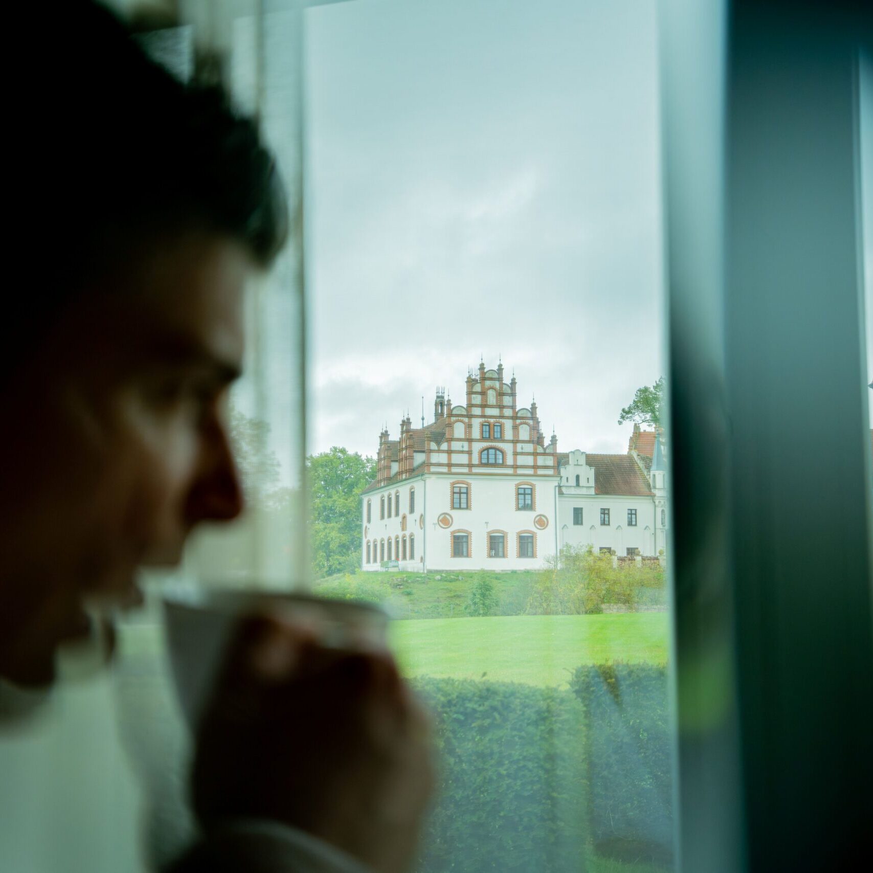Eine Person genießt einen ruhigen Moment, während sie im „Farmer’s Spa“ an einem Fenster aus einer Tasse trinkt; im Glas spiegelt sich ein großes, kunstvoll verziertes Gebäude wider.