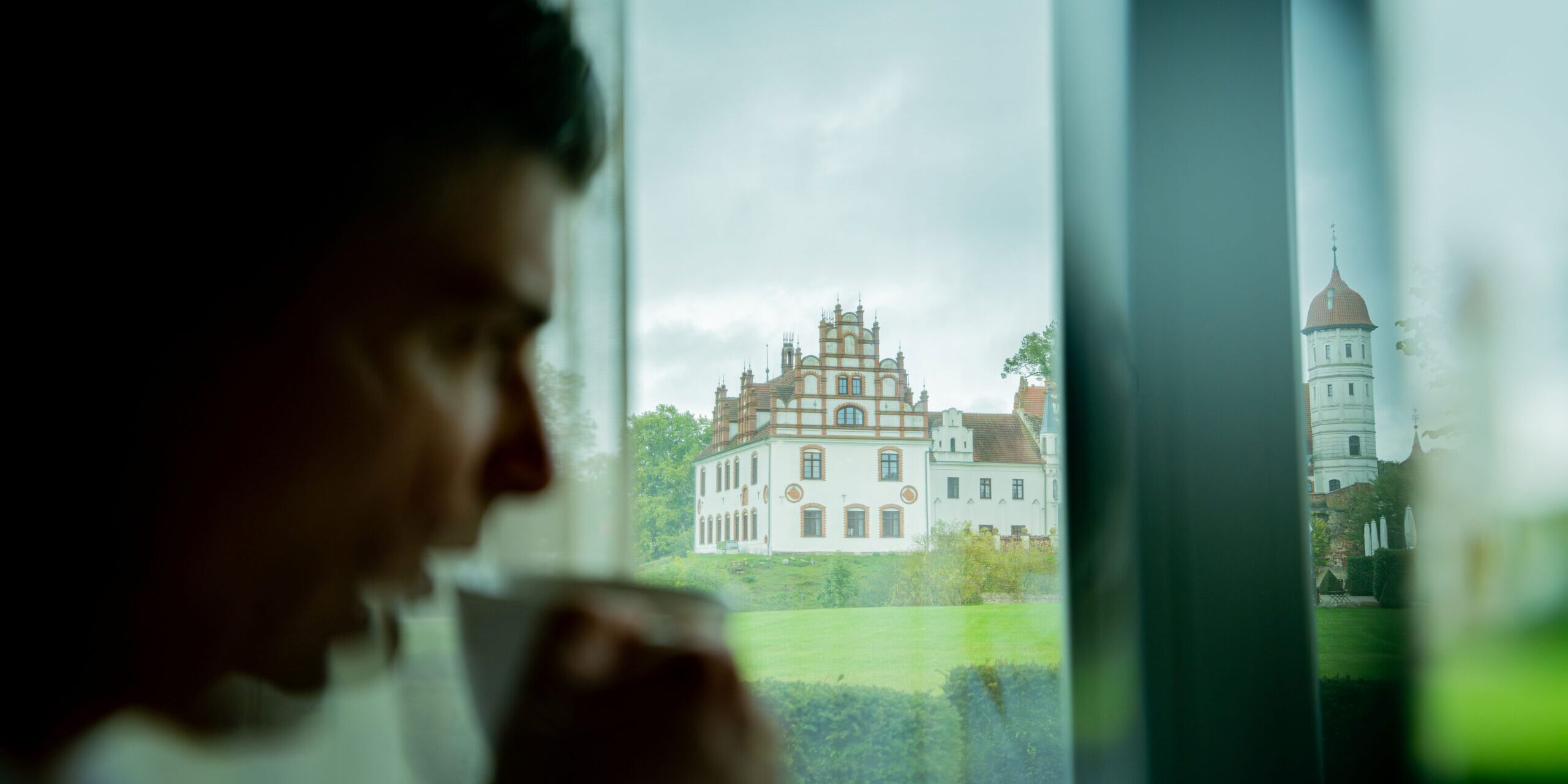 Eine Person genießt einen ruhigen Moment, während sie im „Farmer’s Spa“ an einem Fenster aus einer Tasse trinkt; im Glas spiegelt sich ein großes, kunstvoll verziertes Gebäude wider.