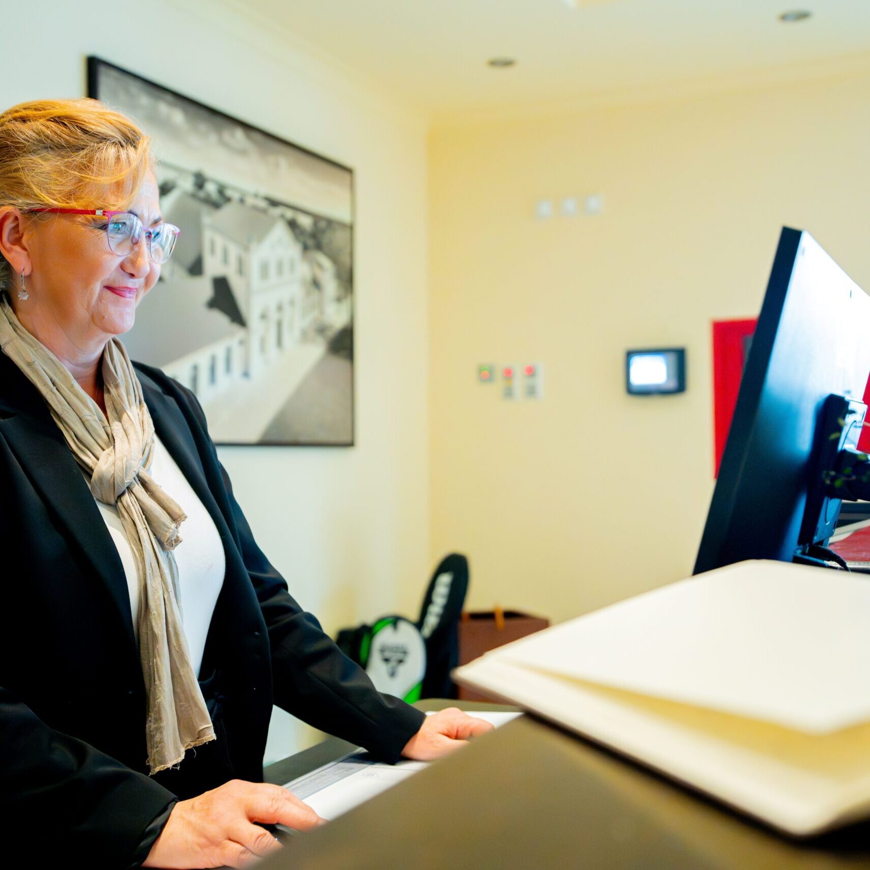 Eine Frau in Business-Kleidung steht an einem Empfangstresen, lächelt, während sie am Computerbildschirm arbeitet, und betreut die Kunden von Sommer & Basedow professionell und herzlich.