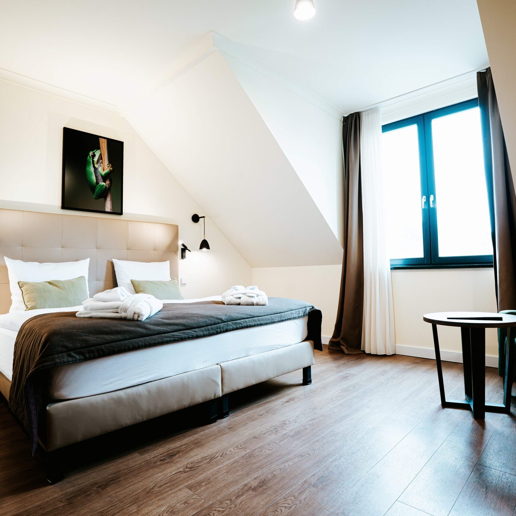 Modernes Hotelzimmer mit Doppelbett, Nachttischlampen, großem Fenster, Holzboden und einer kleinen Sitzecke – ideal zum Entspannen nach einem Tag bei „Golftage“ oder nach einem Ausflug zum nahegelegenen Golfclub Schloss Teschow.